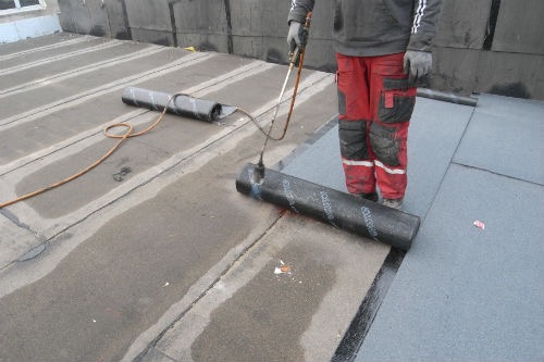 Pracownik w czerwonych spodniach z kieszeniami zgrzewa papę termozgrzewalną na płaskim dachu za pomocą palnika gazowego, widoczne rolki papy i gotowe fragmenty izolacji.