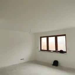 Wnętrze pokoju po malowaniu ścian na biało, z widocznymi puszkami elektrycznymi, brązowym oknem oraz plecakiem leżącym na podłodze.