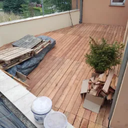 Drewniany taras w trakcie budowy, widoczne deski, palety, donice z kwiatami, wiadro z farbą, oraz mała choinka w doniczce, ujęcie z góry.