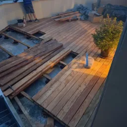 Montaż drewnianego tarasu na balkonie z widocznymi legarami i regulowanymi wspornikami, częściowo ułożone deski, doniczka z iglakiem w rogu.