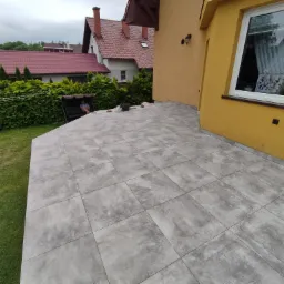 Wyłożony szarymi płytkami taras przy żółtym domu, widok z góry, w tle zieleń i huśtawka z siedzącą osobą.