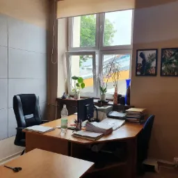 Biuro z biurkiem, krzesłem i komputerem pod oknem, na biurku sterty dokumentów, butelka wody i artykuły biurowe, na ścianie obrazy z motywami roślinnymi.