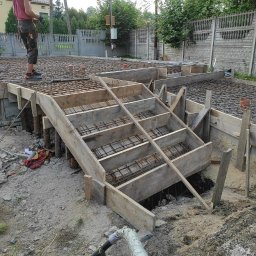 Wood-tech Piotr Gębala - Zbrojenie i szalunek pod fundament płyty, widoczne drewniane schody do wykopu, dwóch pracowników w tle.