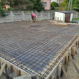 Wood-tech Piotr Gębala - Zbrojenie fundamentów domu jednorodzinnego, widoczne szalunki, siatka z prętów stalowych, folia hydroizolacyjna, pracownicy w tle.