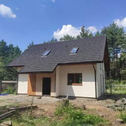 Wood-tech Piotr Gębala - Nowoczesny dom szkieletowy z ciemnym dachem z dwoma oknami dachowymi, zlokalizowany w Olkuszu, otoczony zielenią i drzewami.