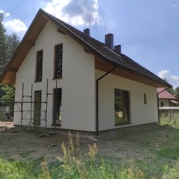 Wood-tech Piotr Gębala - Nowoczesny dom szkieletowy z jasną elewacją, ciemną dachówką i drewnianą podbitką, częściowo otoczony rusztowaniem, w otoczeniu drzew i zieleni.