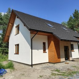 Wood-tech Piotr Gębala - Nowoczesny dom szkieletowy z ciemną dachówką ceramiczną, jasną elewacją i drewnianymi elementami dekoracyjnymi, widok z zewnątrz w słoneczny dzień, niezagospodarowany teren wokół.