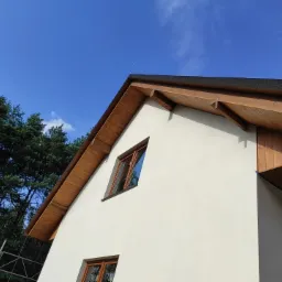 Fragment domu z wykończonym dachem, widoczne drewniane podbitki, okna w brązowych ramach i jasna elewacja na tle błękitnego nieba i zielonych drzew.