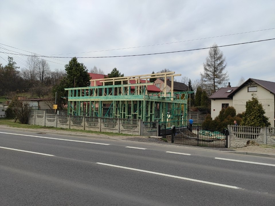 Wznoszenie dwukondygnacyjnej konstrukcji szkieletowej domu z zielonego drewna, widok z ulicy z dwoma pasami ruchu, ogrodzenie betonowe i metalowe, pochmurne niebo.