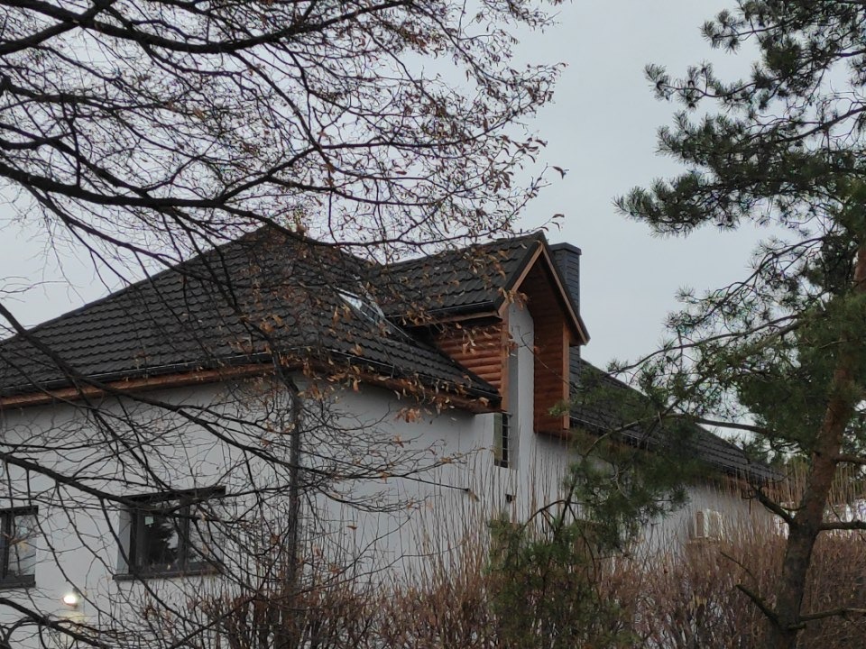Dom jednorodzinny z ciemnobrązowym dachem z dachówki ceramicznej, widoczne okno dachowe i komin, ujęcie z dołu przez gałęzie drzew.