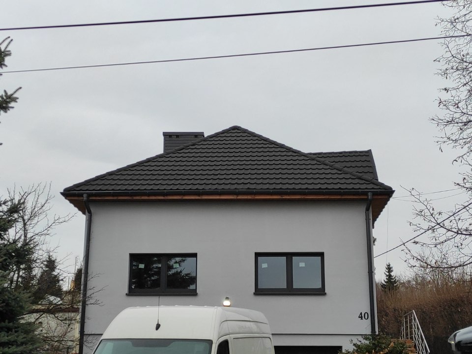 Dom jednorodzinny z nowym, ciemnym dachem z blachodachówki i ciemnymi oknami, widok z dołu, na pierwszym planie biały samochód dostawczy.