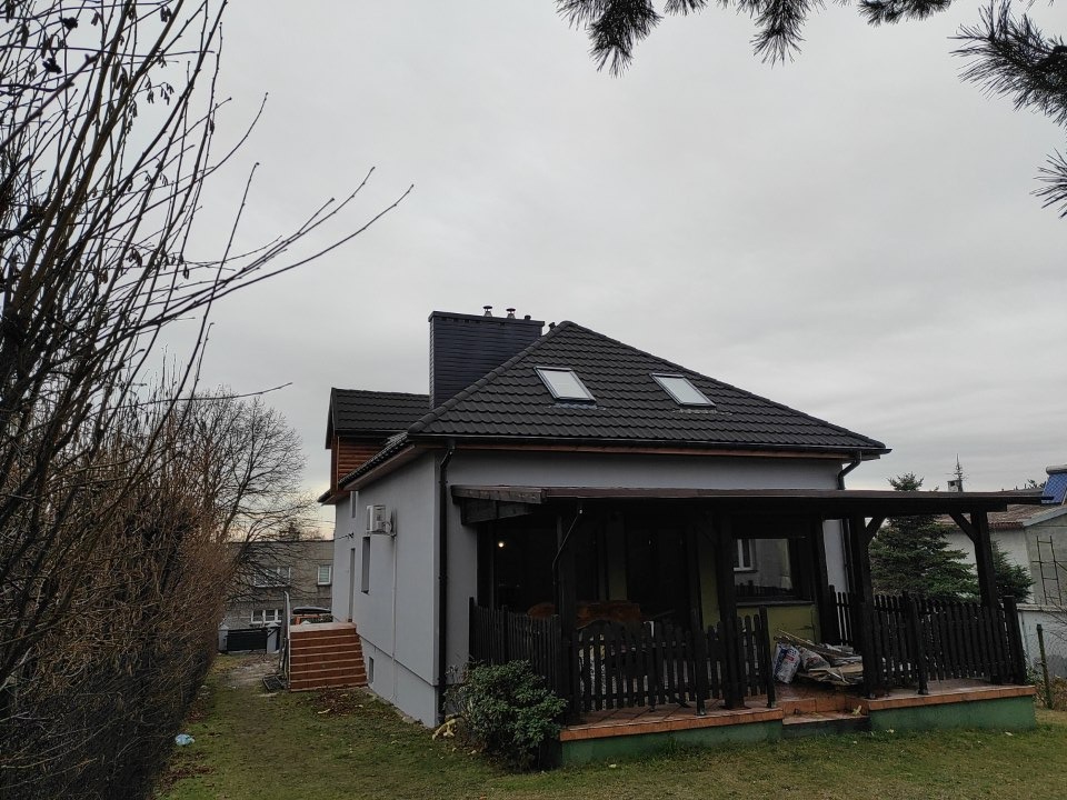 Szary dom jednorodzinny z ciemnym dachem z dachówką i dwoma oknami dachowymi, widok z ogrodu w pochmurny dzień.