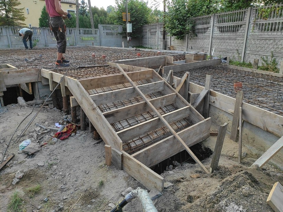 Zbrojenie i szalunek pod fundament płyty, widoczne drewniane schody do wykopu, dwóch pracowników w tle.