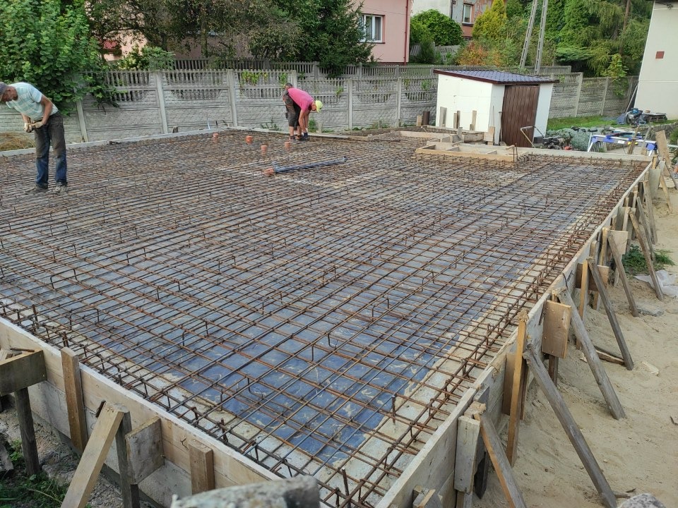 Zbrojenie fundamentów domu jednorodzinnego, widoczne szalunki, siatka z prętów stalowych, folia hydroizolacyjna, pracownicy w tle.