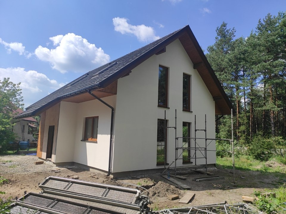 Niedokończony dom szkieletowy z czarną dachówką, widoczne rusztowania i niezagospodarowany teren wokół budynku, w tle las.