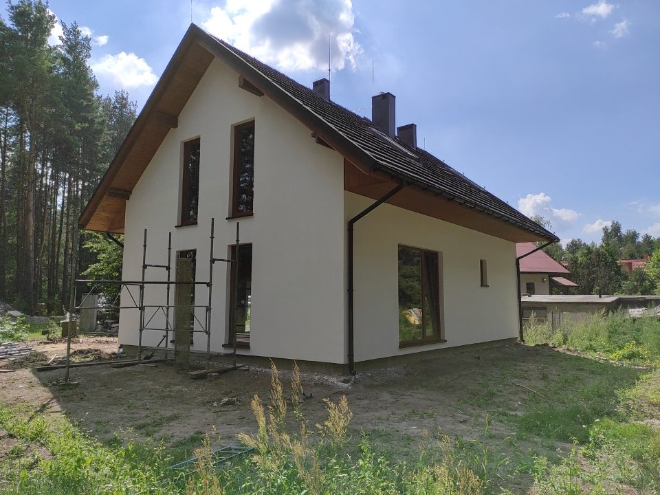 Nowoczesny dom szkieletowy z jasną elewacją, ciemną dachówką i drewnianą podbitką, częściowo otoczony rusztowaniem, w otoczeniu drzew i zieleni.