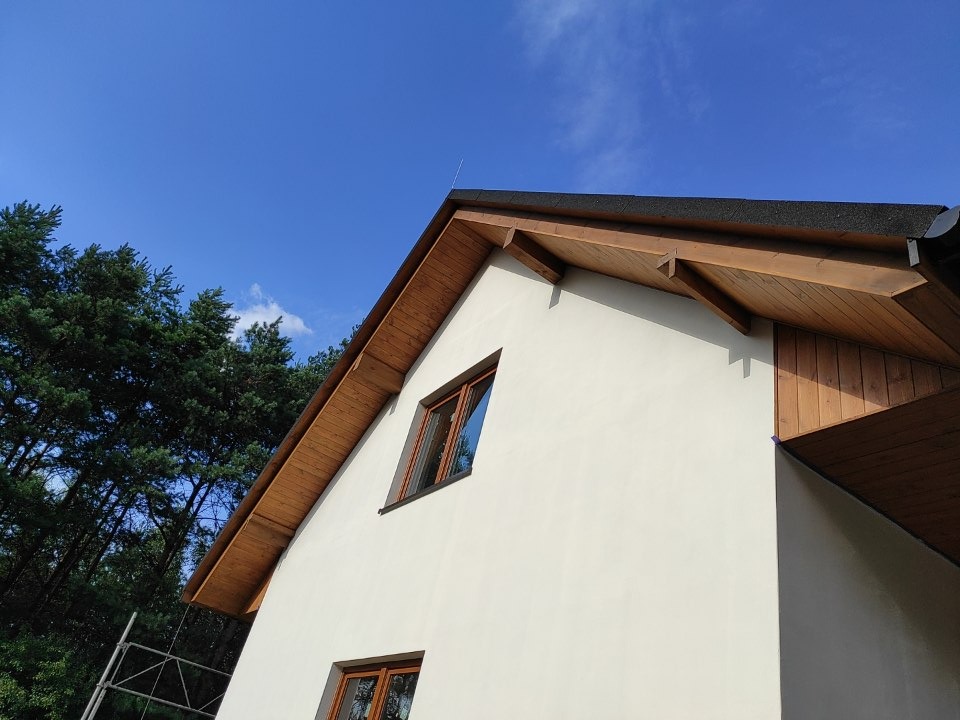 Fragment domu z wykończonym dachem, widoczne drewniane podbitki, okna w brązowych ramach i jasna elewacja na tle błękitnego nieba i zielonych drzew.