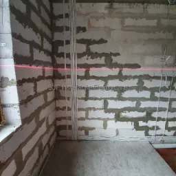 Wnętrze w trakcie remontu z widocznymi przewodami elektrycznymi ułożonymi w korytkach na ścianie z betonu komórkowego, wyznaczonym poziomem lasera i wiertarką akumulatorową na podłodze.