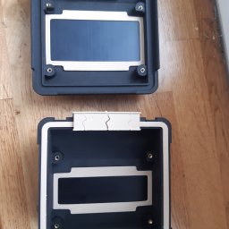 Jakub Szymankiewicz - Dwie czarne, puste obudowy z tworzywa sztucznego, wykonane w technologii druku 3D, z widocznymi warstwami i otworami na śruby, leżące na drewnianym blacie.