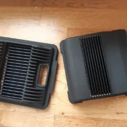 Dwie czarne, plastikowe kratki wentylacyjne z widocznymi żebrami, leżące na drewnianej powierzchni, prawdopodobnie elementy obudowy urządzenia.