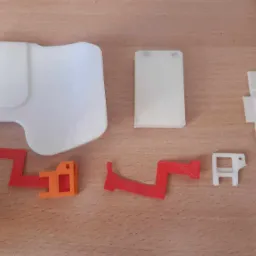 Elementy wydrukowane w 3D w różnych kolorach (biały, czerwony, pomarańczowy) o zróżnicowanych kształtach, leżące na drewnianej powierzchni, widoczne ślady warstw druku.