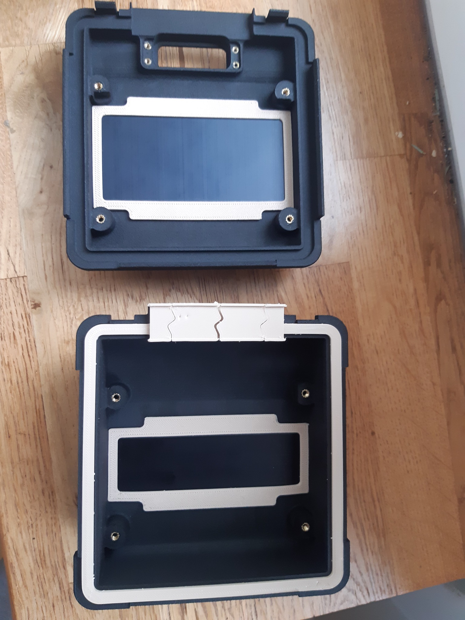 Dwie czarne, puste obudowy z tworzywa sztucznego, wykonane w technologii druku 3D, z widocznymi warstwami i otworami na śruby, leżące na drewnianym blacie.