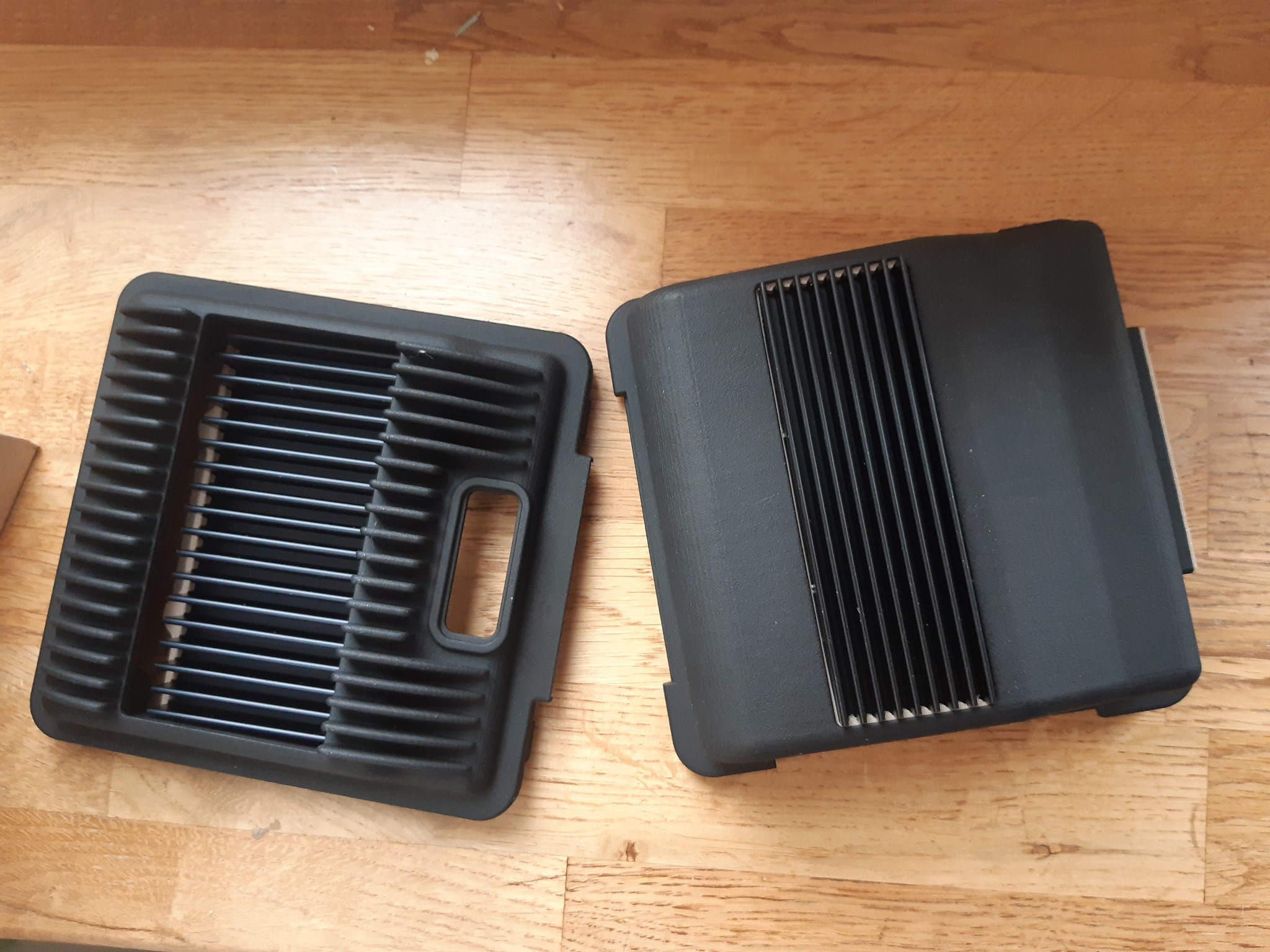 Dwie czarne, plastikowe kratki wentylacyjne z widocznymi żebrami, leżące na drewnianej powierzchni, prawdopodobnie elementy obudowy urządzenia.
