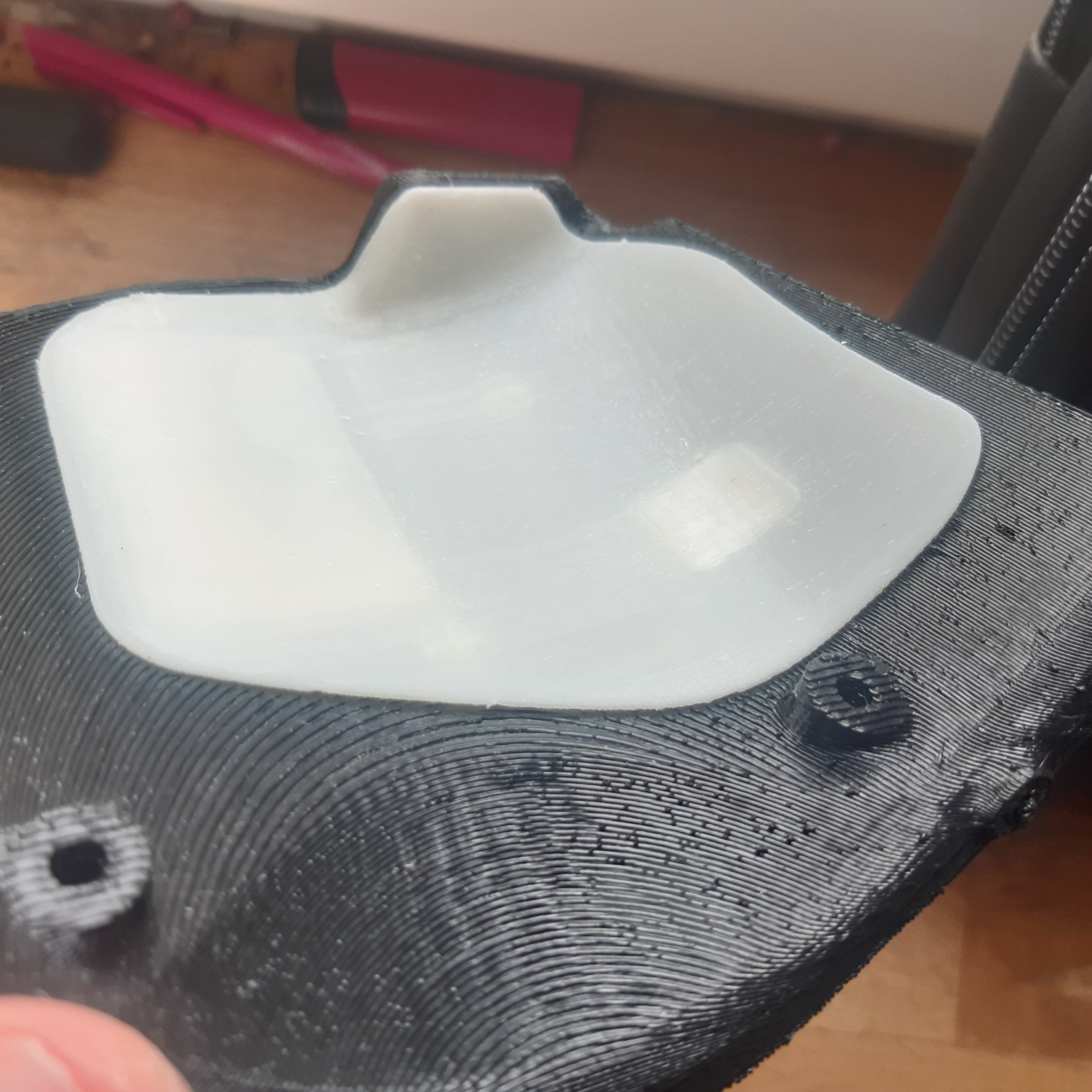 Biały element z tworzywa sztucznego umieszczony na czarnej, drukowanej w 3D podstawie z widocznymi warstwami druku i otworami montażowymi.