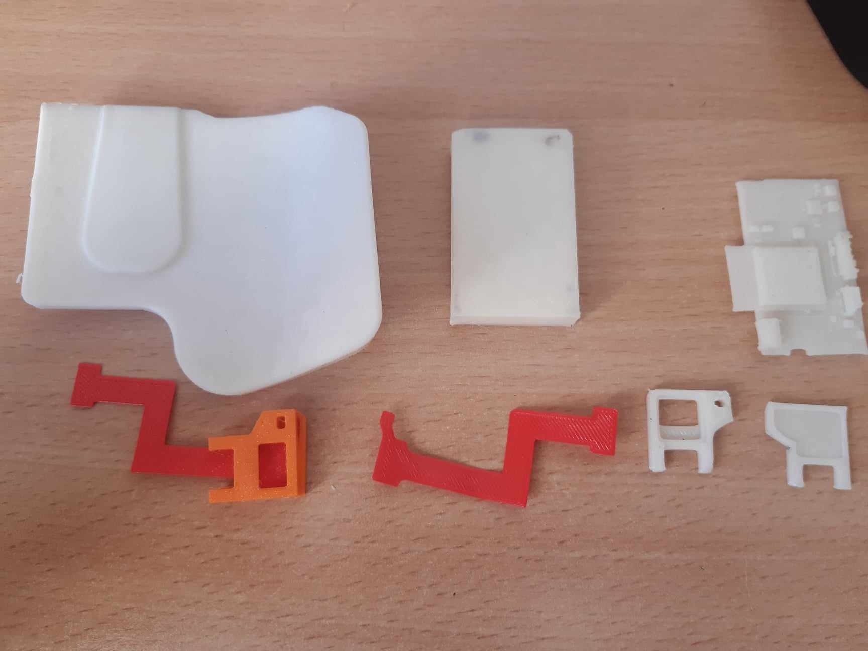 Elementy wydrukowane w 3D w różnych kolorach (biały, czerwony, pomarańczowy) o zróżnicowanych kształtach, leżące na drewnianej powierzchni, widoczne ślady warstw druku.