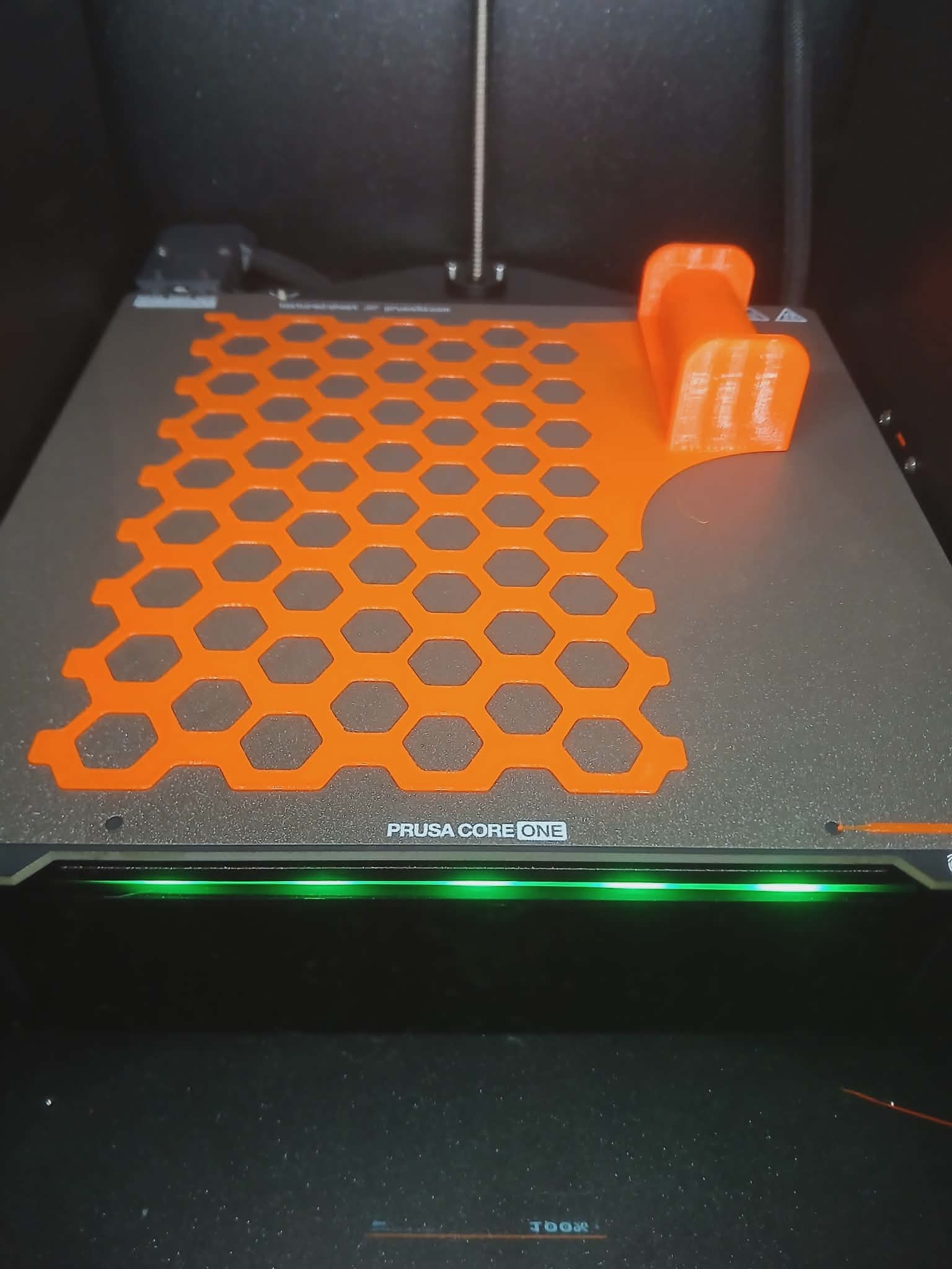 Pomarańczowy wydruk 3D o strukturze plastra miodu i uchwyt na platformie drukarki Prusa Core One, podświetlonej zielonym światłem.