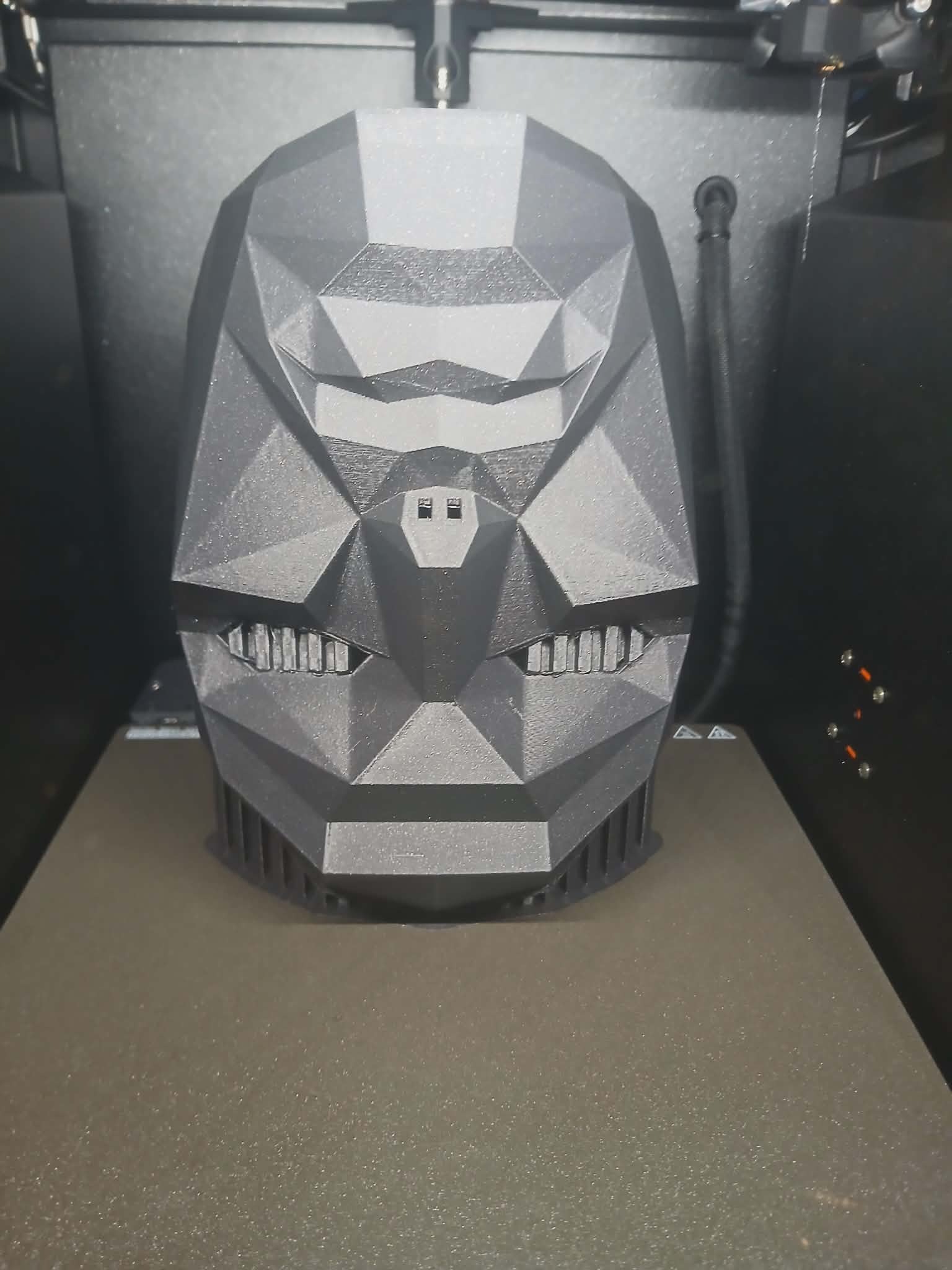 Czarna, geometryczna maska wydrukowana w 3D, umieszczona na platformie drukarki. Widoczne detale konstrukcji i dysza drukująca. Oświetlenie studyjne podkreśla fakturę.