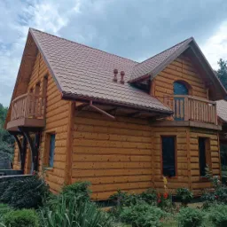 Zewnętrzny widok nowo wybudowanego, dwukondygnacyjnego domu z bali z brązowym metalowym dachem i drewnianymi balkonami, okna zabezpieczone folią.