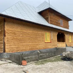 Drewniany dom po renowacji z nowym metalowym dachem, okna zabezpieczone folią malarską, widoczny fragment ogrodu i kamienne schody wejściowe.