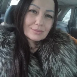 Portret kobiety w futrze z długimi, ciemnymi włosami, siedzącej w samochodzie.