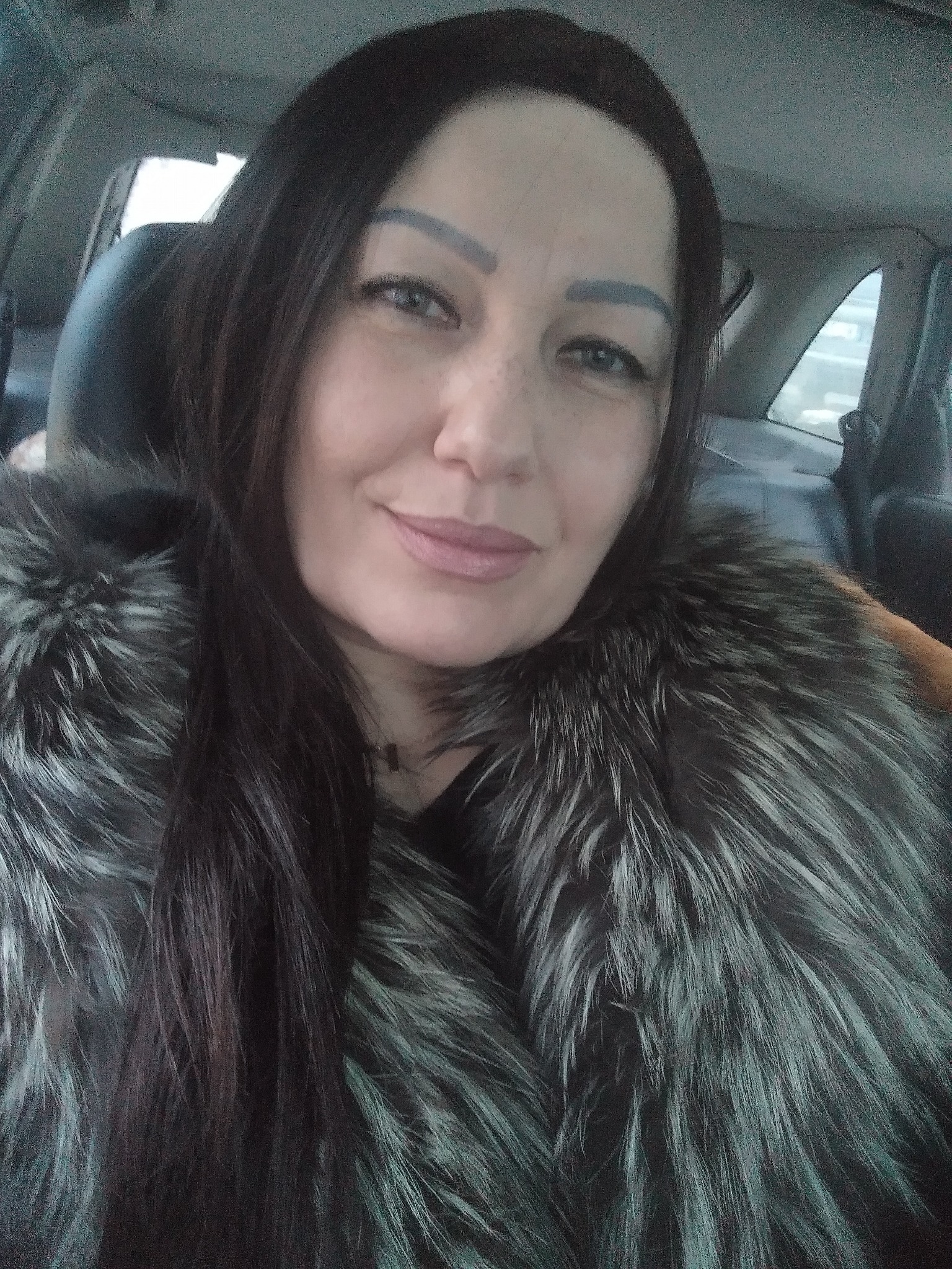 Portret kobiety w futrze z długimi, ciemnymi włosami, siedzącej w samochodzie.
