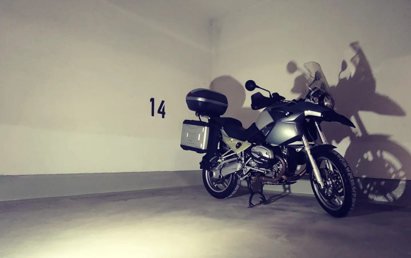 Motocykl turystyczny BMW z kuframi bocznymi i centralnym, zaparkowany w garażu oznaczonym numerem 14, rzucający cień na ścianę.