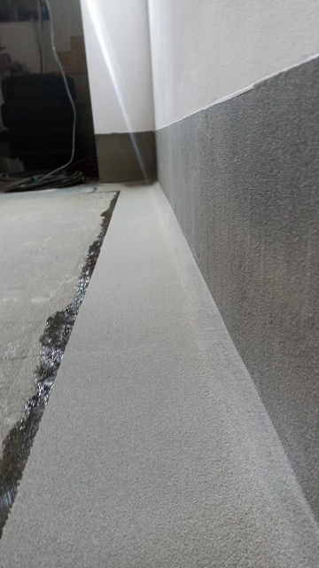 Świeżo wylana, jasnoszara posadzka w rogu pomieszczenia, z widocznymi śladami wilgoci przy ścianie i ciemniejszą, chropowatą listwą przypodłogową.
