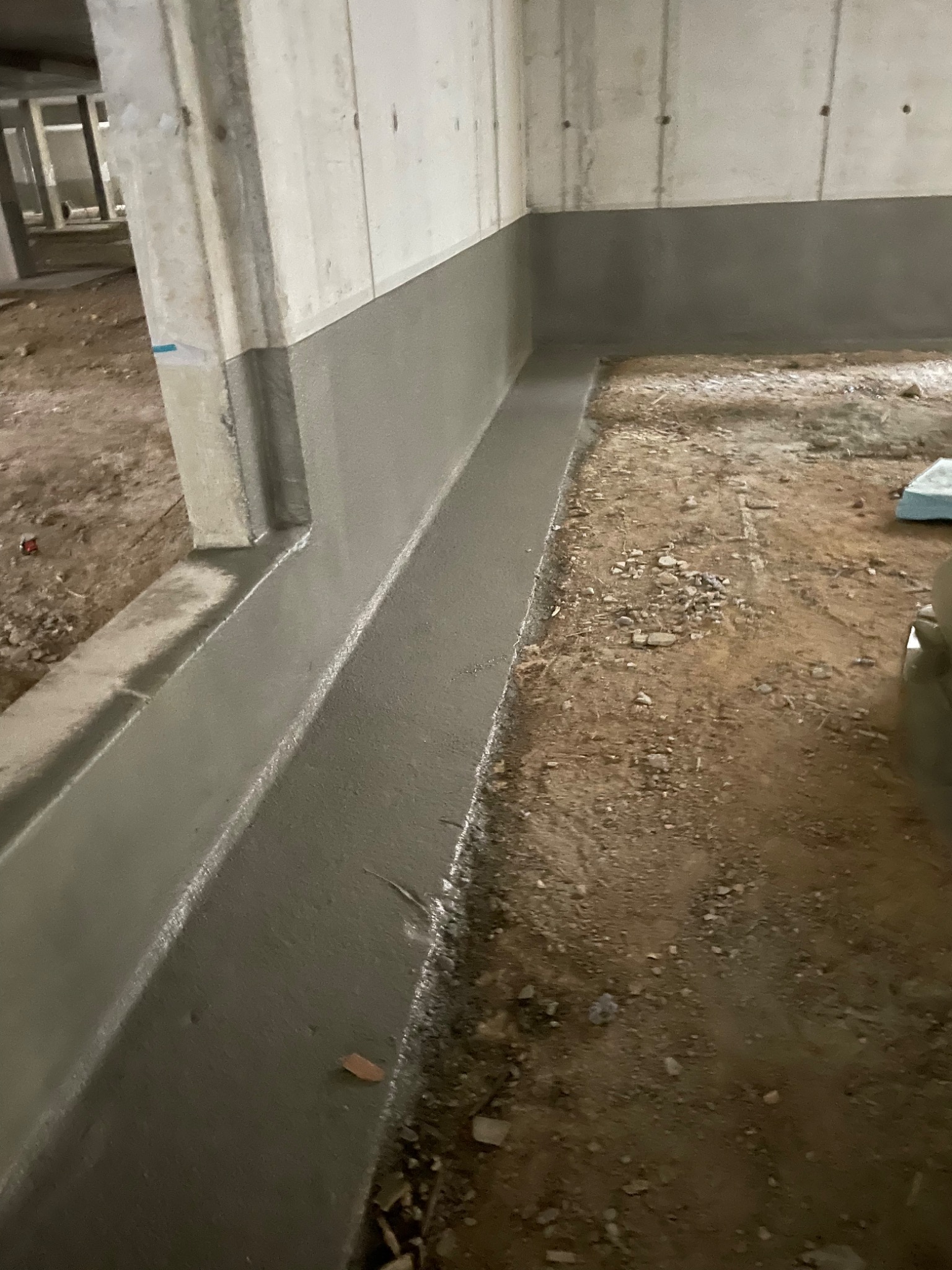 Świeżo wylana, gładka posadzka jastrychowa przy betonowej ścianie w surowym wnętrzu, widoczne niedoskonałości podłoża.