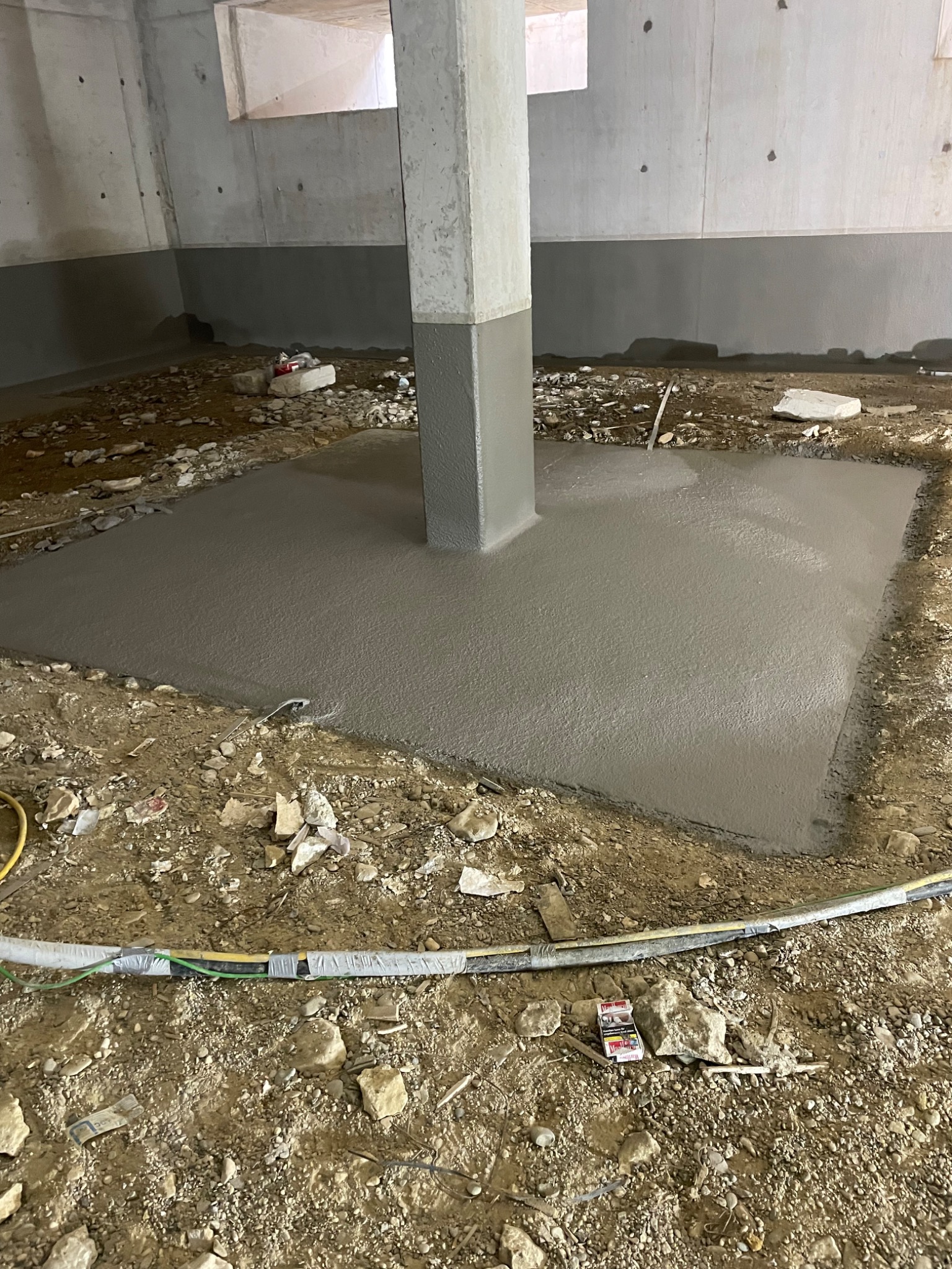 Świeżo wylana, gładka wylewka betonowa wokół betonowego filaru w surowym wnętrzu budynku, widoczne kable i drobne gruz na ziemi.