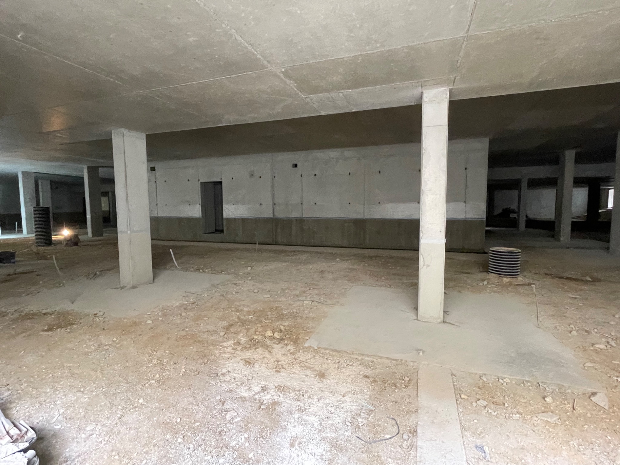 Surowe wnętrze w trakcie prac budowlanych: betonowe filary, ściany i podłoże z widocznymi elementami konstrukcyjnymi. W tle widoczne otwory drzwiowe i instalacje.