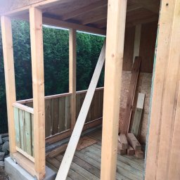 Vitalik Kovalchuk - Wnętrze altany w trakcie budowy: surowe drewno, podłoga z desek, widoczne elementy konstrukcyjne i deski opierające się o filar.