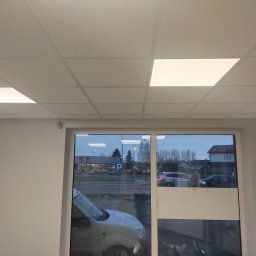Vitalik Kovalchuk - Biały sufit podwieszany z kwadratowymi panelami oświetleniowymi LED, widok przez okno na ulicę z zaparkowanymi samochodami i budynkami w tle.