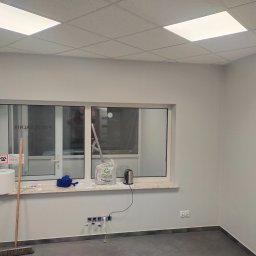 Vitalik Kovalchuk - Świeżo pomalowane na szaro ściany pomieszczenia z oknem, widoczny parapet z przedmiotami, podłoga z ciemnymi panelami i gniazdka elektryczne na ścianie.