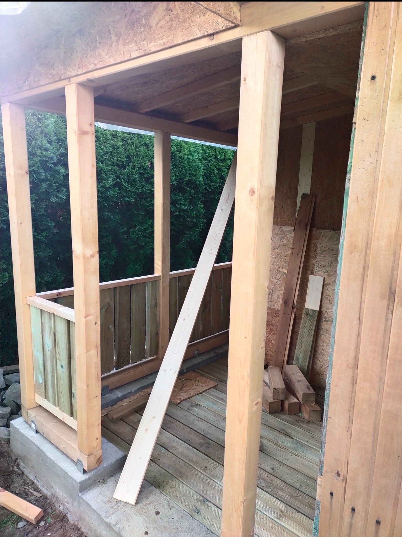 Wnętrze altany w trakcie budowy: surowe drewno, podłoga z desek, widoczne elementy konstrukcyjne i deski opierające się o filar.