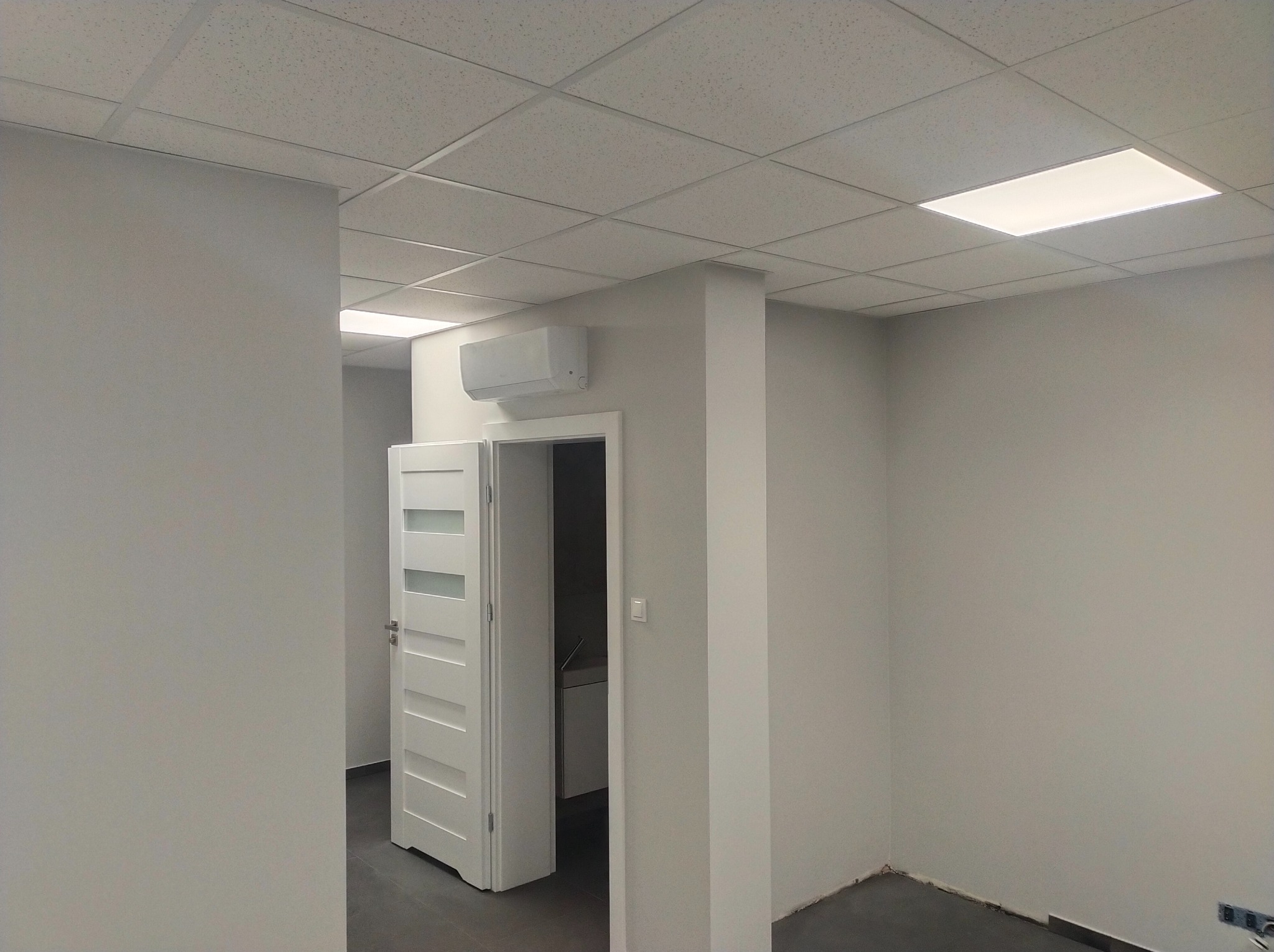 Wnętrze biurowe po odświeżeniu: pomalowane ściany w odcieniach szarości, białe drzwi z przeszkleniami, sufit podwieszany z oświetleniem LED i klimatyzator.