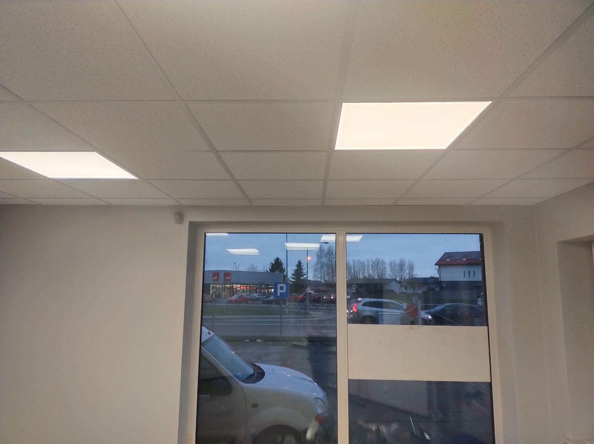 Biały sufit podwieszany z kwadratowymi panelami oświetleniowymi LED, widok przez okno na ulicę z zaparkowanymi samochodami i budynkami w tle.