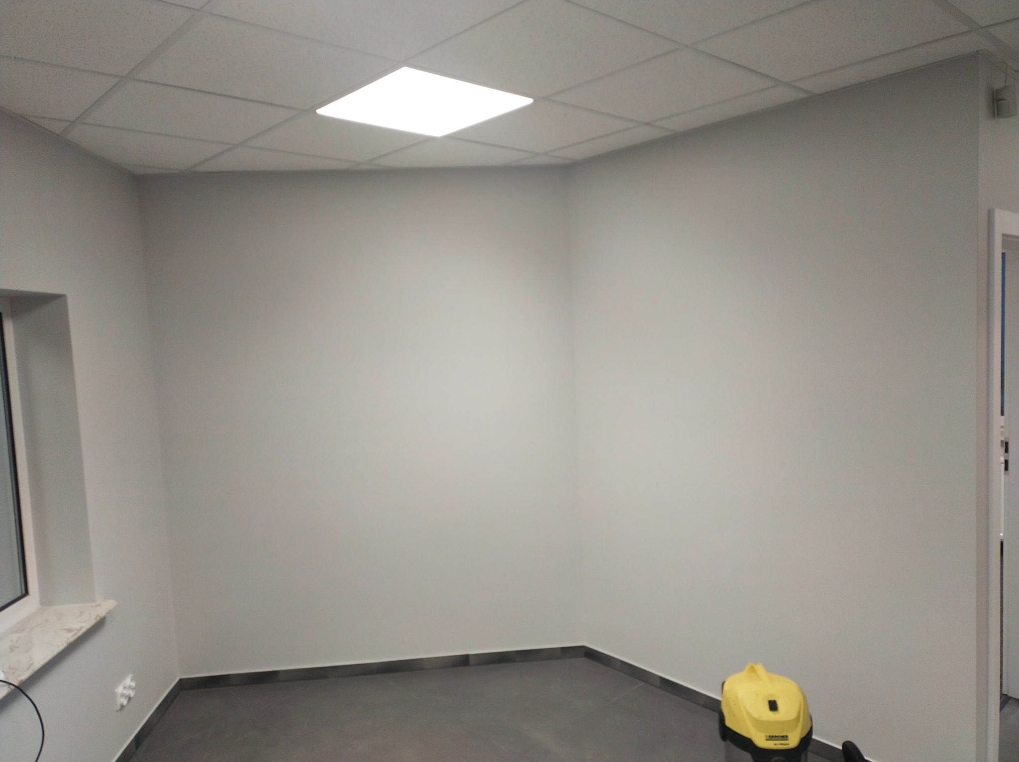 Pomieszczenie po malowaniu ścian na szaro, widoczny szary panel podłogowy i odkurzacz przemysłowy Karcher w rogu.