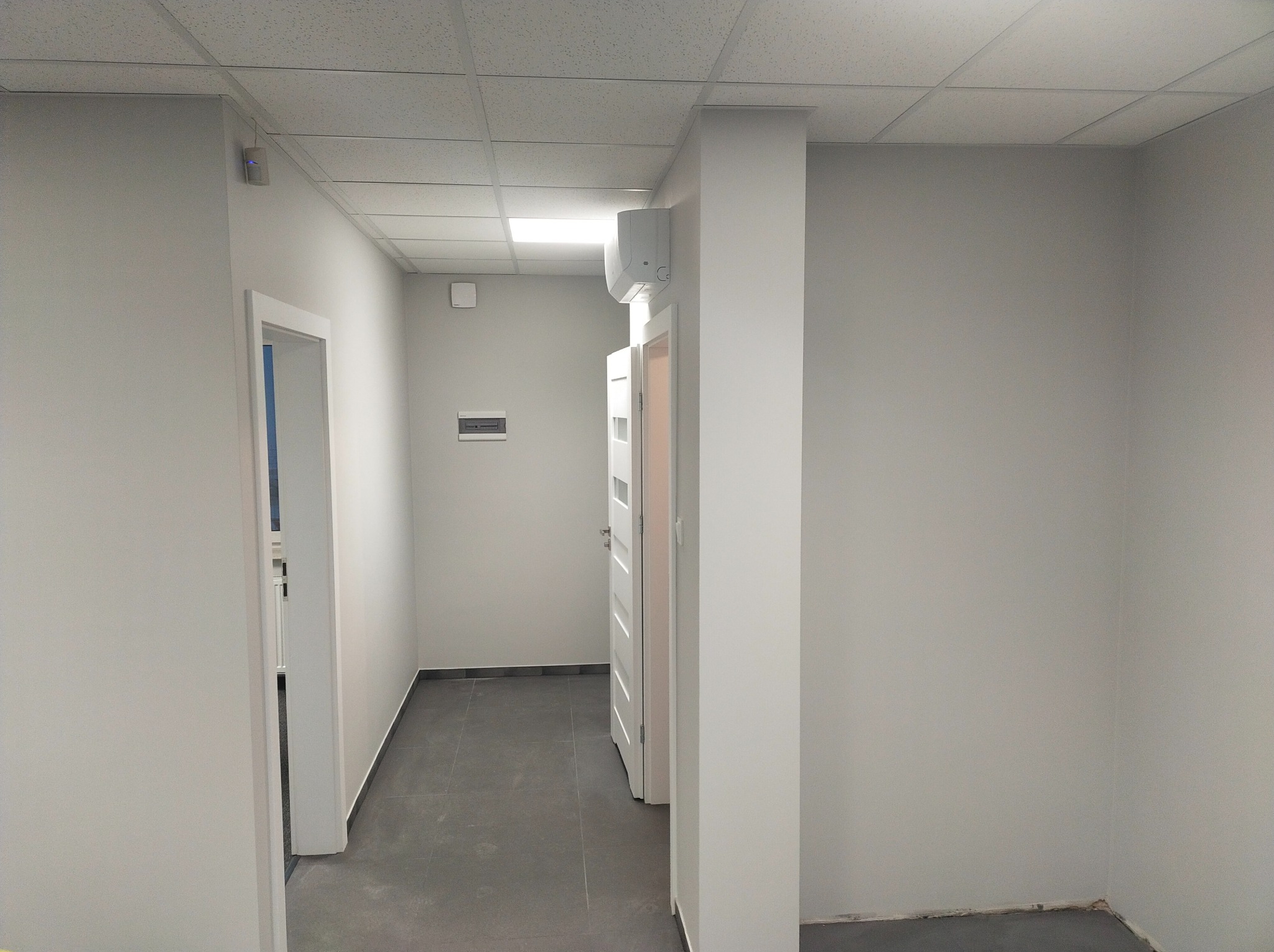 Jasnoszare wnętrze korytarza z białymi drzwiami, szarą podłogą i klimatyzacją na ścianie, widok po odświeżeniu.