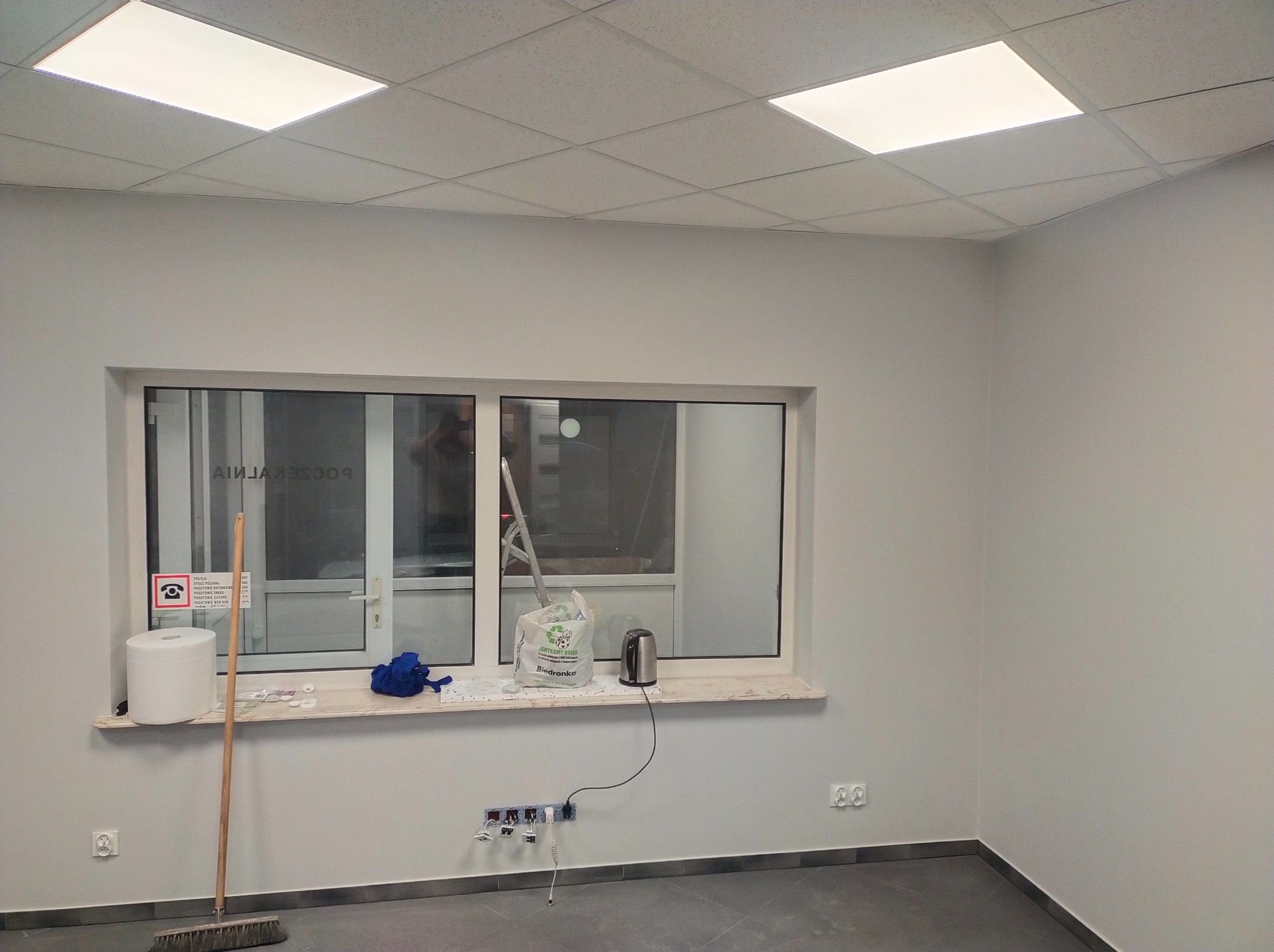 Świeżo pomalowane na szaro ściany pomieszczenia z oknem, widoczny parapet z przedmiotami, podłoga z ciemnymi panelami i gniazdka elektryczne na ścianie.