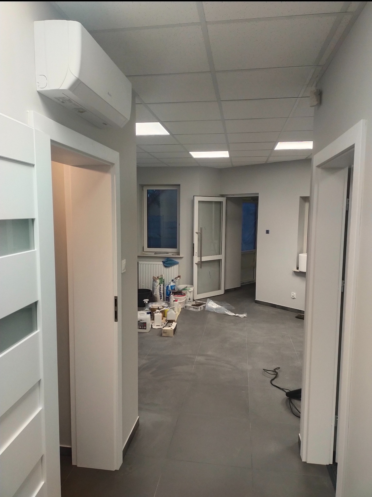 Wnętrze biurowe w trakcie prac wykończeniowych; widoczne białe ściany, szara podłoga, klimatyzator na ścianie, otwarte drzwi, okno z grzejnikiem oraz materiały budowlane.